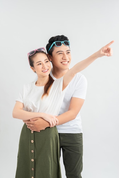 Happy asian man hugging smiling girlfriend while pointing with finger isolated on white