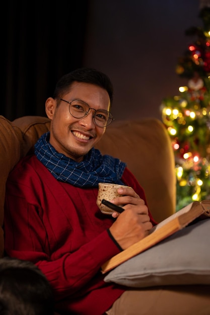 A happy Asian man is relaxing on a couch on Christmas night sipping hot cocoa and reading a book