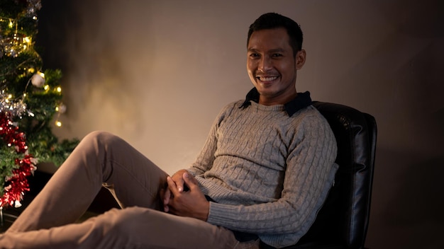 A happy Asian man is sitting on a chair near by a Christmas tree in his dark living room