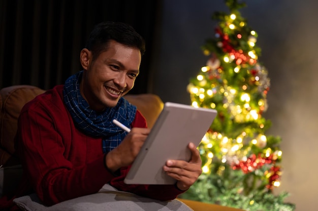 A happy Asian man is using his digital tablet on a couch in his living room on a Christmas night