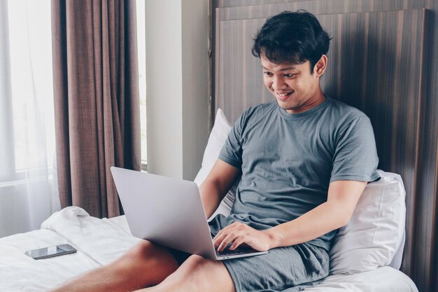 Happy Asian man is working with his laptop on his bed Concept of freelancer successful lifestyle