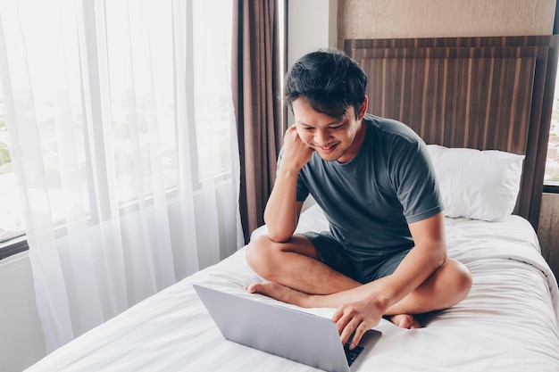 L'uomo asiatico felice sta lavorando con il suo laptop sul suo letto concetto di stile di vita di successo del libero professionista