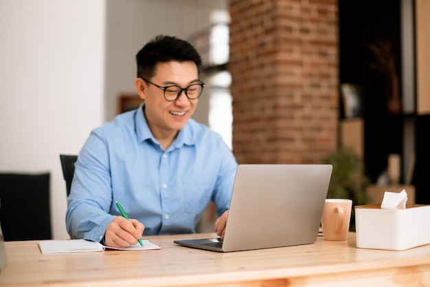 행복한 아시아 남자가 노트북 화면을 보고 홈오피스 선별적 초점에 있는 직장에 앉아 노트북에 글을 씁니다.