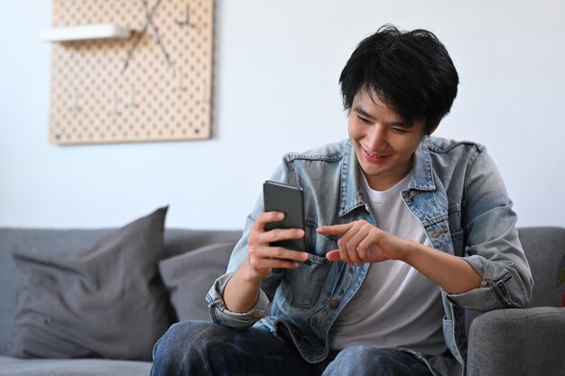彼のスマートフォンでソーシャルメディアをチェックしてオンラインで買い物をする幸せなアジア人男性