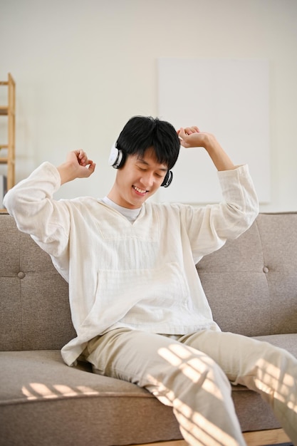 Happy Asian man sits in sofa and enjoys listening to music through headphones