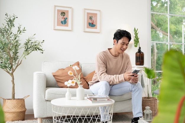 A Happy asian man sitting on couch using smartphone in living room at home