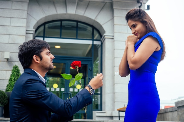 Happy asian man in stylish black suit falling knee in front of his beautiful woman in blue long dress asking proposal and giving gold ring on the background of the restaurant street.