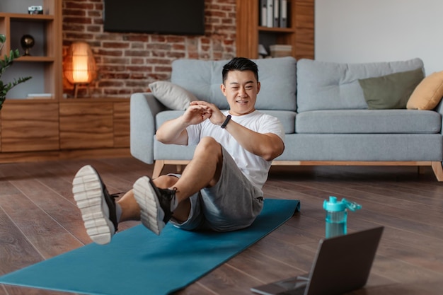 Happy asian middle aged man doing exercises for legs and abs on mat and watching workout on computer