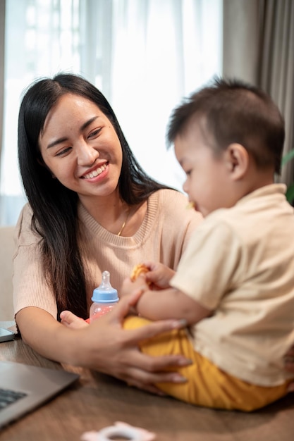 A happy Asian mom is playing and feeding her little son with snacks while working in her home office