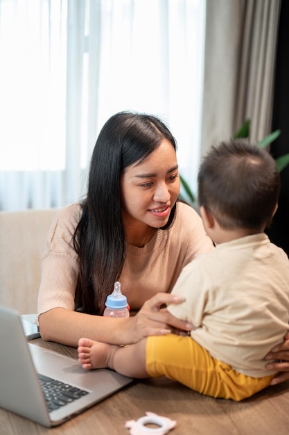 幸せなアジアの母親がラップトップで働きながら小さな息子と遊んでいます