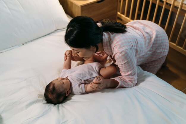 happy asian mother is leaning over and playing with her cute baby daughter near the bed before bedtime at nighttime in the bedroom at home.