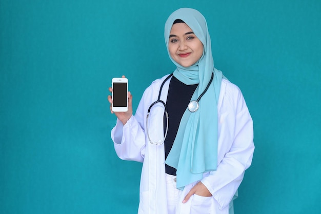 Happy Asian muslim doctor or nurse with stethoscope smiling and showing smartphone over green backgr