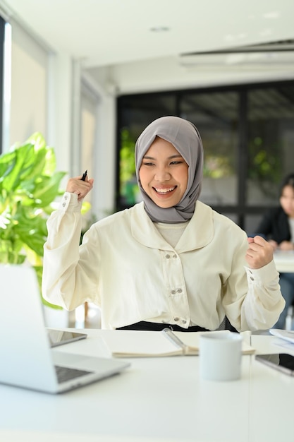 Счастливая азиатская офисная работница-мусульманка поднимает руки вверх, празднуя свой успешный проект