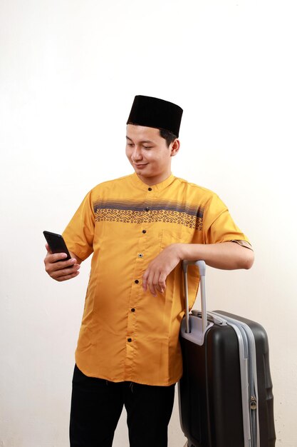 Photo happy asian muslim man carrying travel suitcase while holding a cell phone going mudik or travel to hometown for ramadan eid mubarak celebration