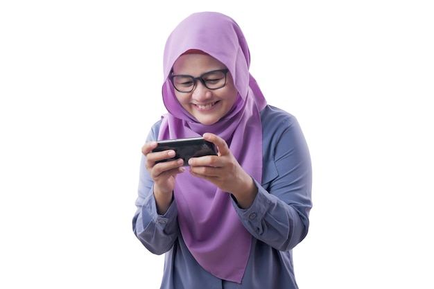 Счастливая азиатская мусульманка с удовольствием играет в игры на своем смартфоне изолирована на белом