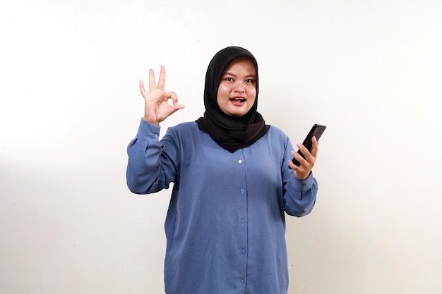 Happy asian muslim woman holding a cell phone while making okay hand gesture