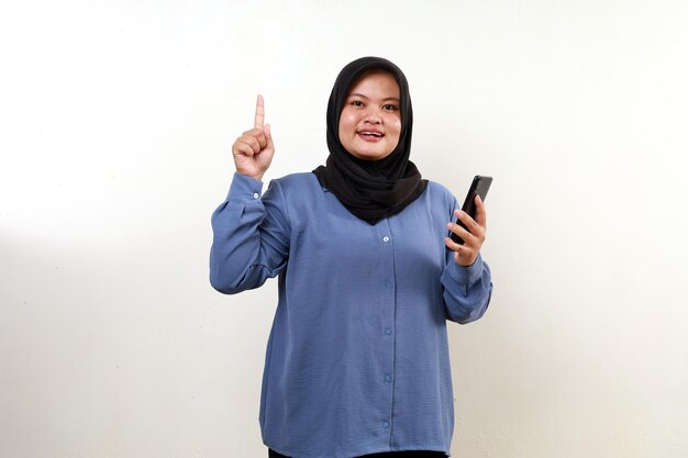 Happy asian muslim woman holding a cell phone while pointing above