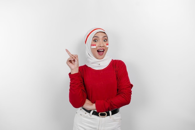 A happy Asian muslim woman wearing white hijab and red top and pointing copy space on top of her isolated by white background Indonesia's independence day