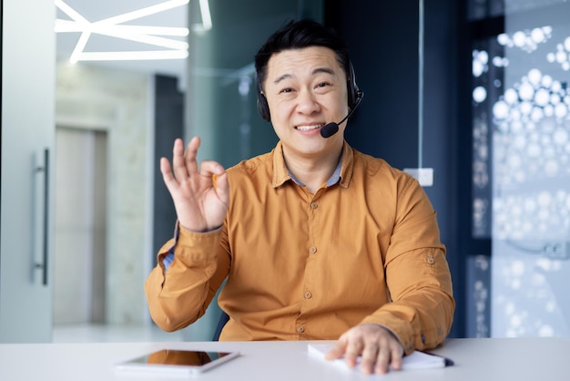 Happy asian office worker sitting at desk in bright office wearing headset and talking on video call
