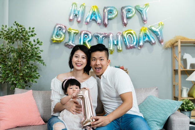 happy asian parents looking camera while celebrating first birthday of their baby daughter who’s playing with the number standee at home with colorful decoration