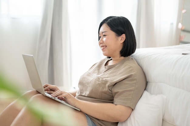 幸せなアジア人のプラスサイズの女性が、寝室でリラックスしながら自宅で仕事をしながらラップトップを使用しています