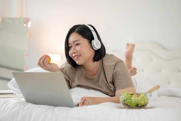 A happy Asian plussize woman lying on bed watching a movie on bed reading an online blog