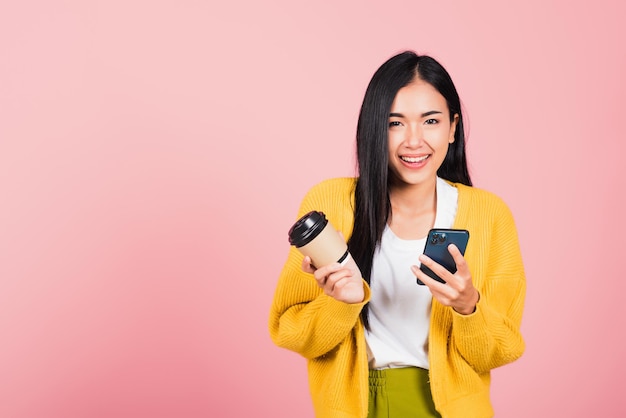 Happy Asian portrait beautiful cute young woman excited smiling holding mobile phone and coffee to go, studio shot isolated on pink background, female using smartphone with coffee cup take away