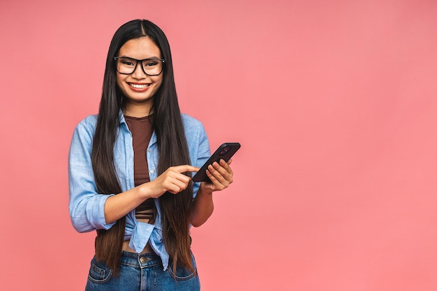 Happy Asian portrait beautiful cute young woman teen smiling excited typing text message on smart mobile phone enjoys online communication isolated on pink background with copy space