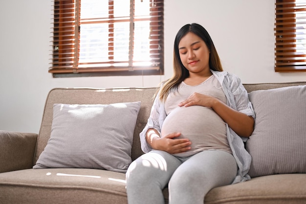 Happy Asian pregnant woman in comfortable clothes relaxes on the sofa in her living room