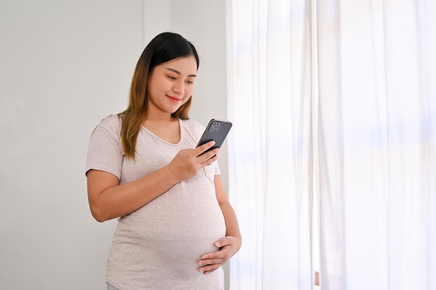 Happy Asian pregnant woman stands by the window using her smartphone and touching her belly