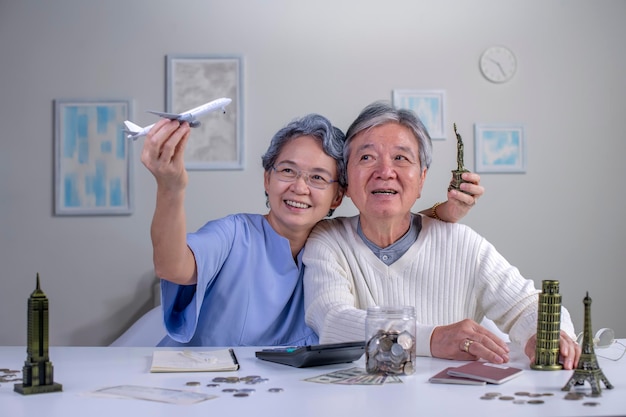 Happy asian senior couple plans to save money for a trip around\
the world travel budget concept passport monument model and\
aircraft toy in the picture