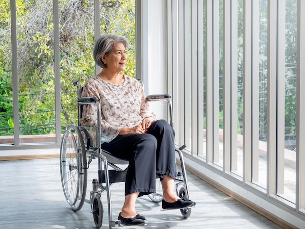 Happy Asian senior woman in casual dress sits in wheelchair smile and looking out of the glass window with natural green view Positive thinking leads to better health in the elderly