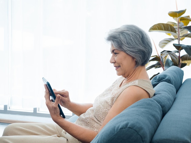 Счастливая азиатская пожилая женщина сидит на диване, держа и глядя на экран цифрового планшета в руке в гостиной