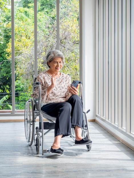 Happy Asian senior woman sits in wheelchair waving greetings smile and looking at smart mobile phone screen vertical style Elderly females meet family via video call near glass window green view