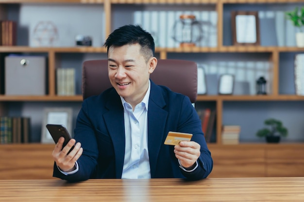 Happy Asian shopping online sitting in the office man using phone app and bank card businessman enjoying online shopping