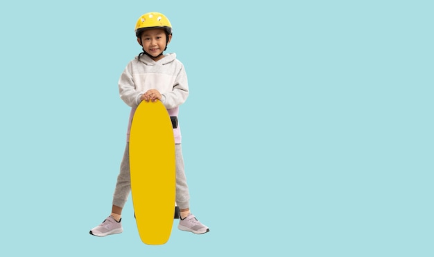 Счастливая азиатская улыбающаяся маленькая девочка, играющая на скейтборде в шлеме Портрет всего тела на пастельном простом голубом фоне