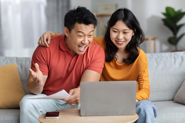 Happy asian spouses reading papers using laptop at home