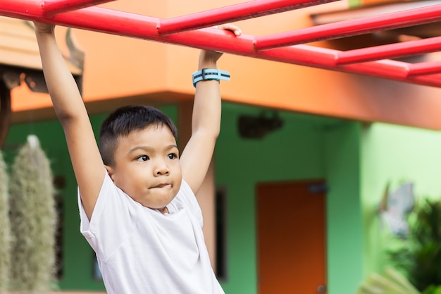 遊んで、遊び場で棒鋼からぶら下がっている幸せなアジア学生子少年。