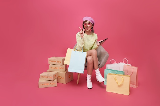 Happy Asian teen woman sitting on sofa holding shopping bags and smartphone