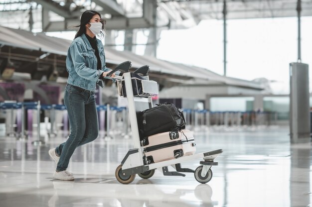 Счастливая азиатская туристка с защитной маской от коронавируса гуляет с тележкой для багажа