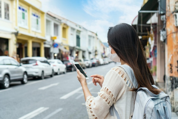 プーケットタイの旧市街を歩き、携帯電話を使用して医療マスクを身に着けている幸せなアジアの旅行者の女性コロナウイルスパンデミックの新しい通常のライフスタイルの概念の間に旅行