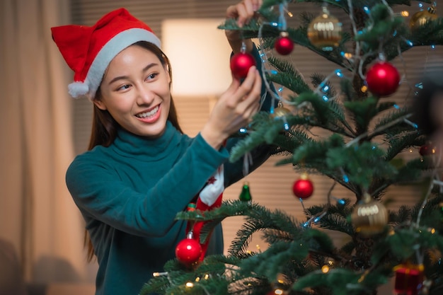 幸せなアジアの女性はメリークリスマスの前にクリスマスツリーを屋内で飾る