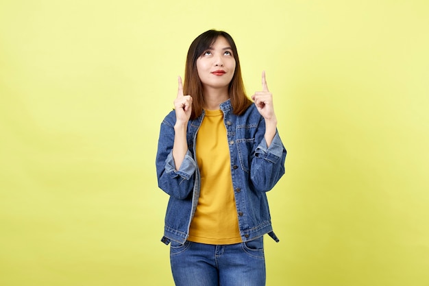 Happy asian woman in denim jacket pointing up to copy space and looking at the camera over yellow space