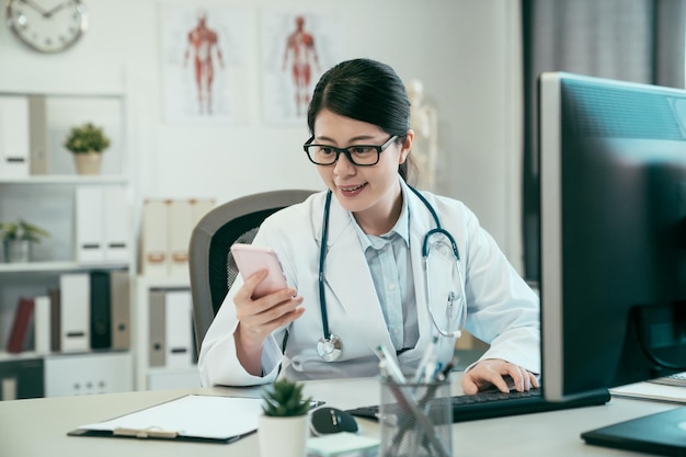 Medico asiatico felice della donna che utilizza smartphone mentre lavora con il personal computer nell'ospedale dell'ufficio. giovane e bello personale medico professionale sorridente leggendo un messaggio di testo sul telefono cellulare in clinica.