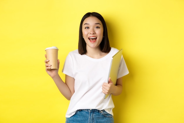 Happy asian woman drinking coffee and holding laptop, saying hi to you, standing over yellow background