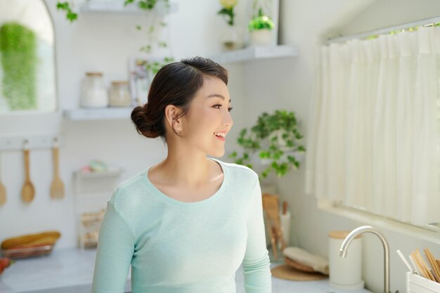 Happy asian woman drinking milk and looking away while standing in kitchen