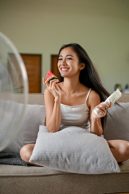 A happy Asian woman enjoys eating fresh watermelon while using an electric handy fan on a couch