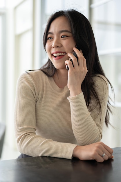 A happy Asian woman enjoys talking on the phone while sitting at a table indoors