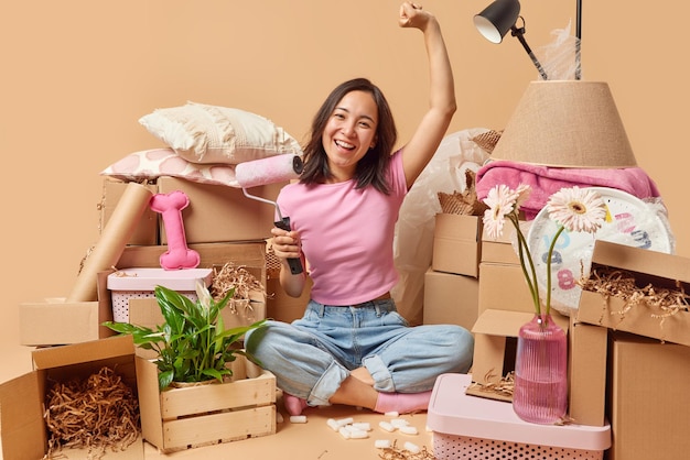 Happy asian woman going to paint walls in room relocates to new\
apartment dressed casually sits in lotus pose surrounded by stacks\
of cardboard boxes golds paint roller house improvement\
concept