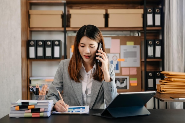 幸せなアジアの女性は、現代のオフィスでスマートフォンのタブレットとラップトップで話す喜びを持っています xA